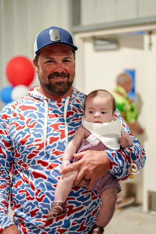 Man in blue cap and patterned shirt holding a baby indoors, smiling at a festive event