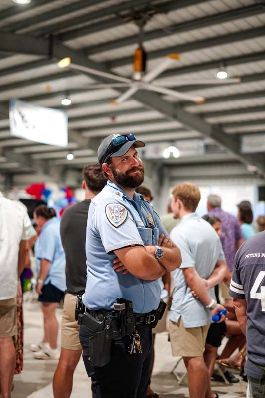 Security guard standing with arms crossed in a busy indoor event hall, wearing a light blue uniform and cap.
