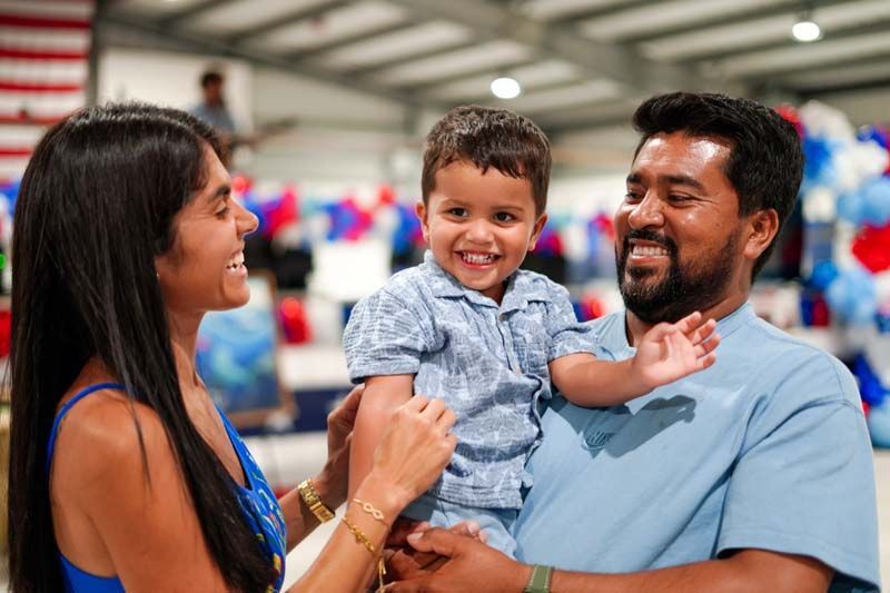 Smiling family holding a young child at an indoor event, with colorful booths in the background