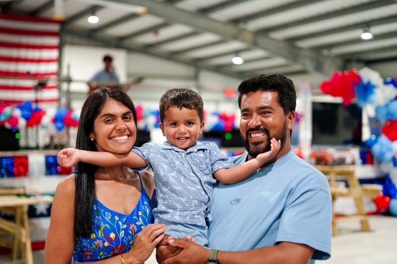 Smiling family posing indoors with a child between two adults, red-white-blue decorations in the background