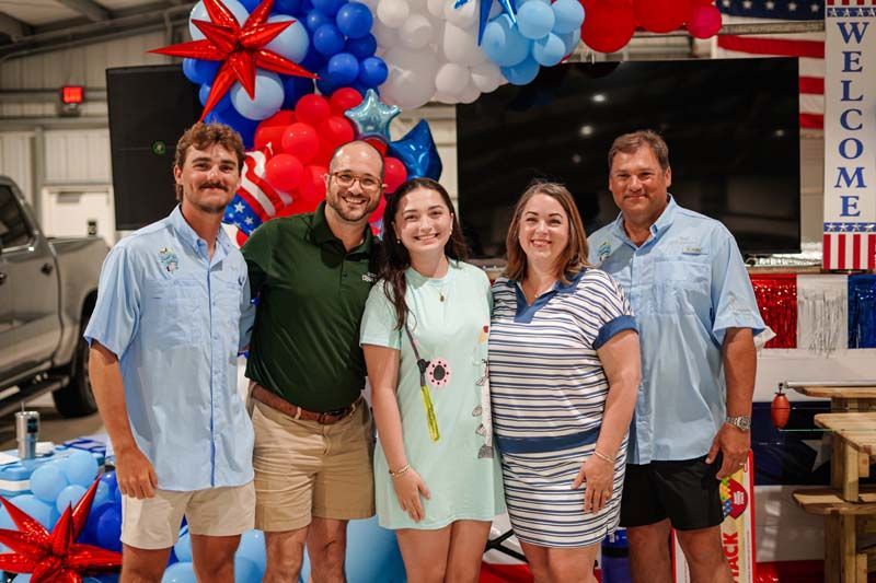 Five people posing at a festive vehicle dealership event with red, white, and blue balloons and a “Welcome” sign