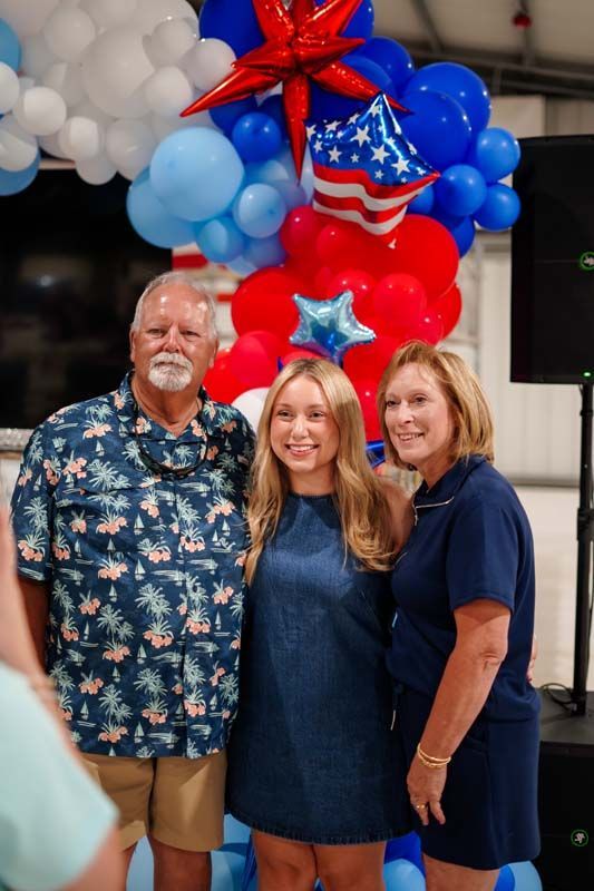 Three people posing indoors beneath red, blue, and white balloon decorations