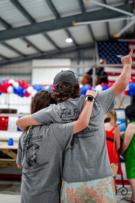 Three people in gray shirts hug at an indoor event with red, white, and blue decorations.