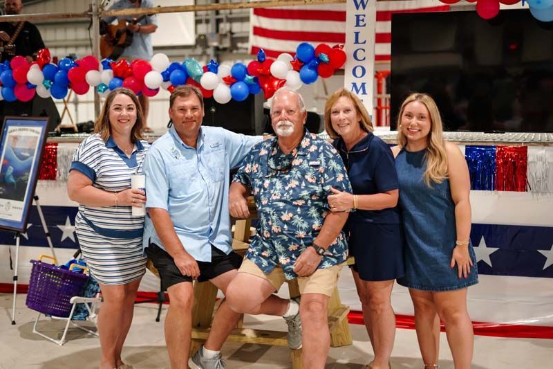 Five people posing in front of red, white, and blue bunting and a “WELCOME” sign at an event