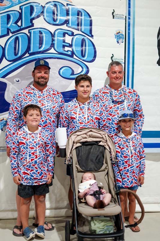 Family posing in matching red, white, and blue shirts in front of a rodeo sign, with a stroller in front
