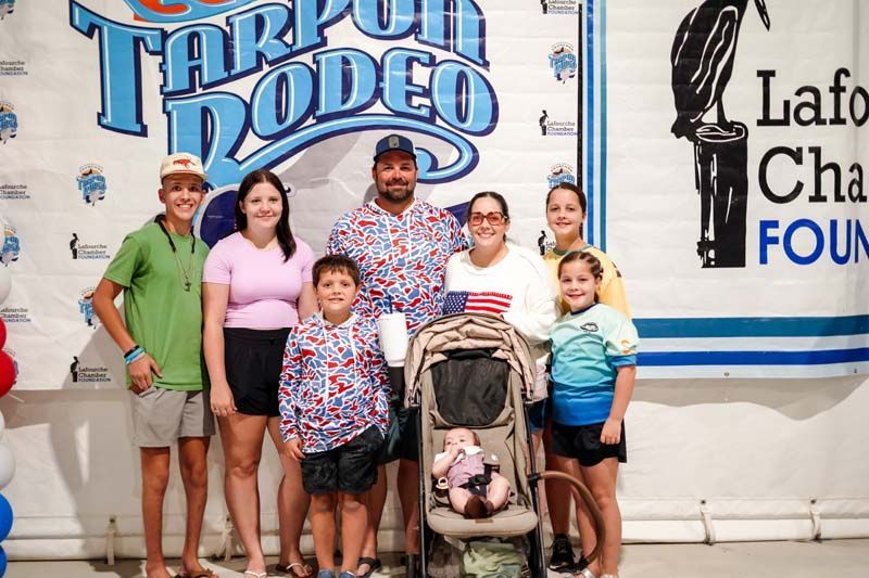 Group of people posing in front of a Tarpon Rodeo sign, with a stroller and wall signage behind them.