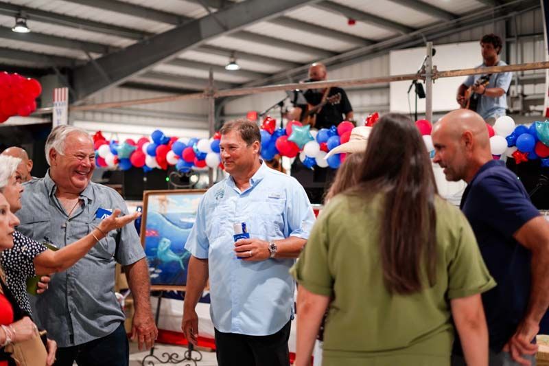 People talking at an indoor event with red, white, and blue balloons and posters in the background
