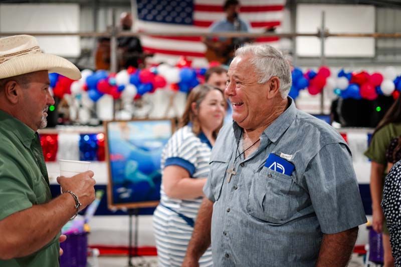 Two men talking at a patriotic indoor event with red, white, and blue balloons and an American flag backdrop