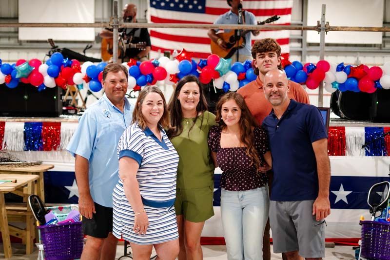 Six people posing in a decorated hall with red, white, and blue balloons and an American flag backdrop