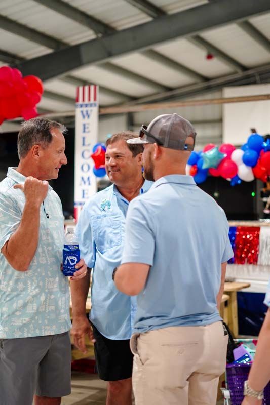 Three men talk at an indoor event with red, white, and blue balloons and a “WELCOME” banner.