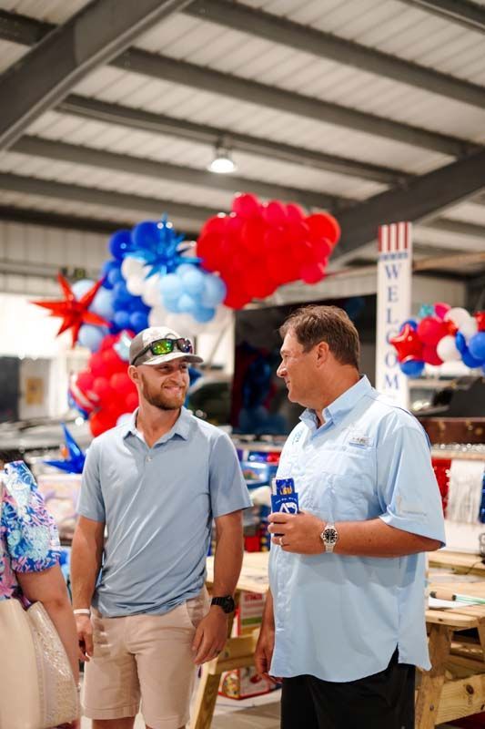 Two men talking at a patriotic indoor event with red, white, and blue balloons and beer cases nearby
