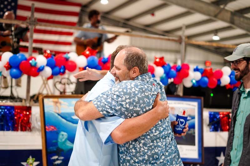 Two men hugging at a patriotic indoor event with red, white, and blue balloons and an American flag backdrop