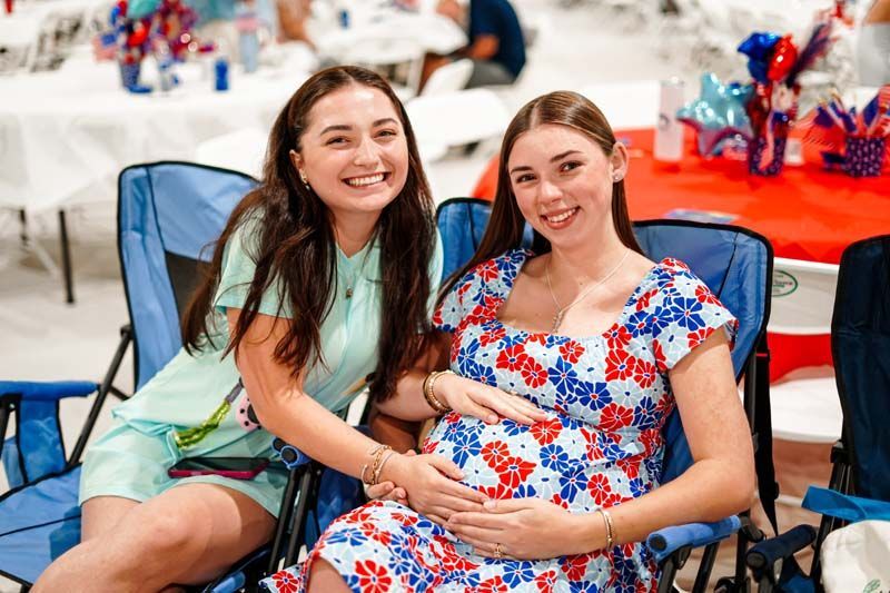 Two women smiling and seated at a decorated indoor event, one in a mint dress and one in a floral dress