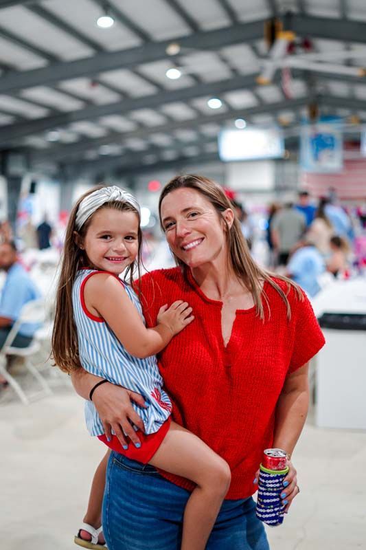 Woman in a red top holding a smiling child in a blue dress at an indoor event hall