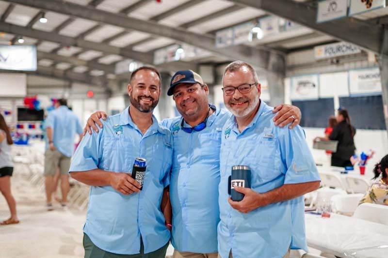 Three men in blue shirts smiling and holding drinks at an indoor event hall