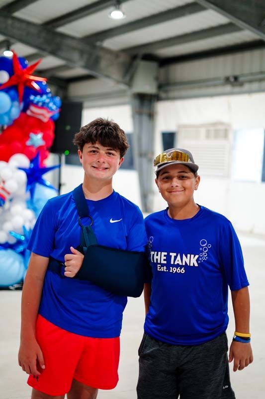 Two people smiling indoors, one with an arm in a sling, standing beside red, white, and blue balloons.
