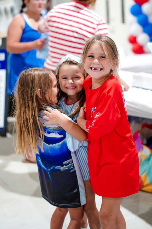 Three children smiling and hugging at a party with red, white, and blue decorations