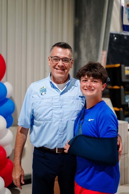 Two smiling people standing indoors, one with an arm in a sling, in front of red and blue balloons.