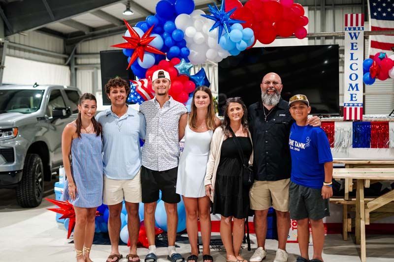 Group posing indoors under red, white, and blue balloons beside a “Welcome” sign.