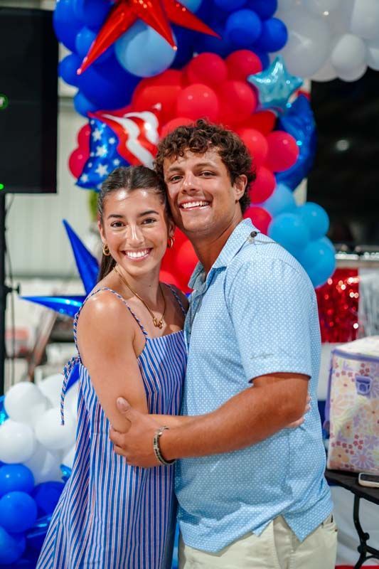 Couple posing in front of red, blue, and white balloon decorations, smiling at the camera.