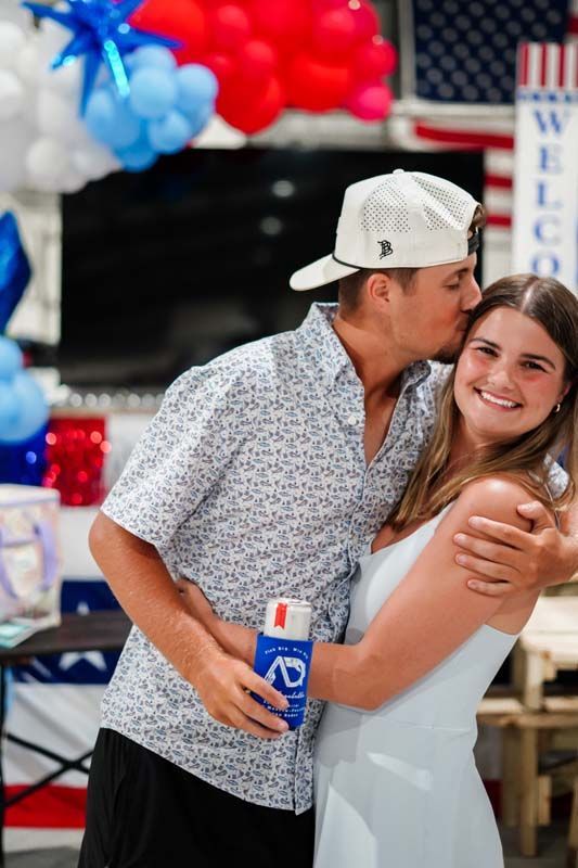 Person in a white cap kisses a smiling woman holding a Bud Light can at a festive event.