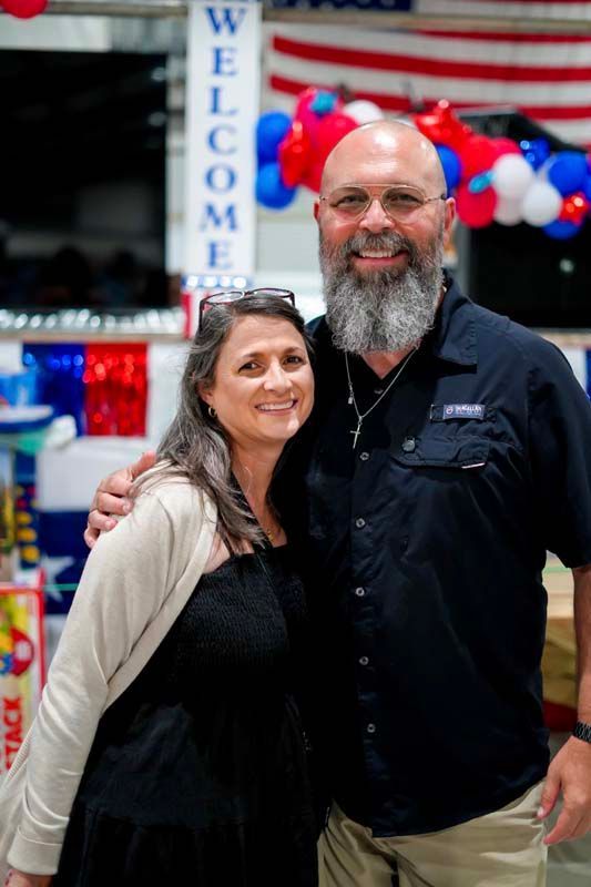 Smiling couple posing at a patriotic indoor event with balloons and a “WELCOME” banner.