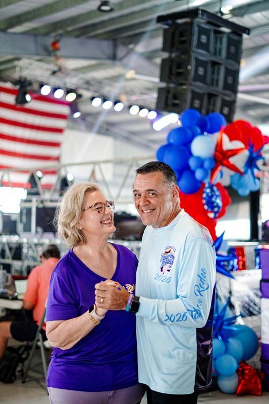 Smiling pair holding hands at a patriotic indoor event with red, white, and blue balloons and an American flag.