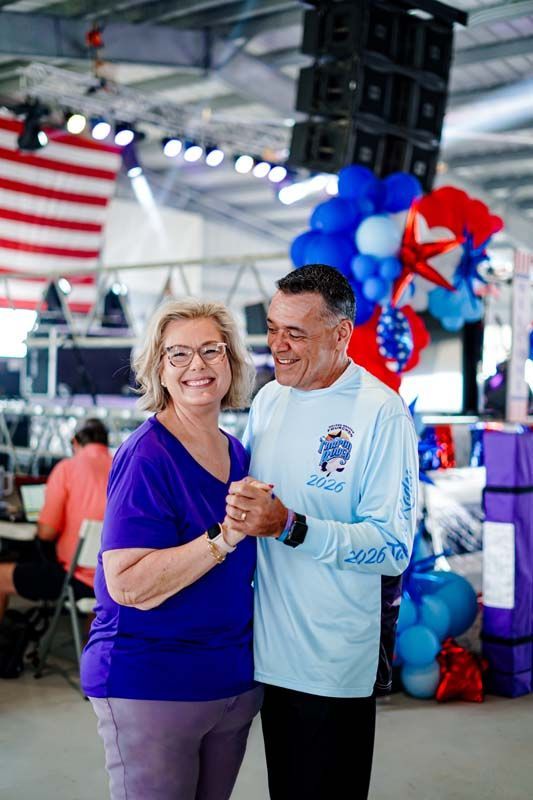 Smiling couple holding hands at an indoor event with blue and red balloons and an American flag backdrop