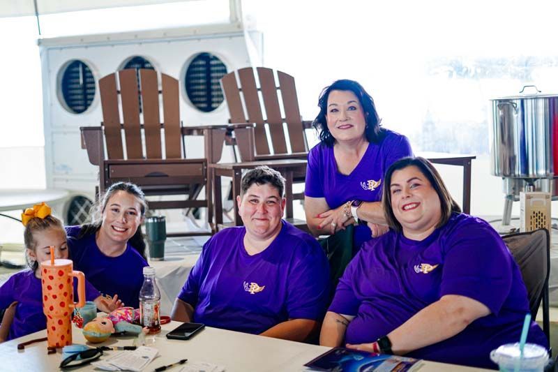 Five people in purple shirts sit at a table with craft supplies in a bright room