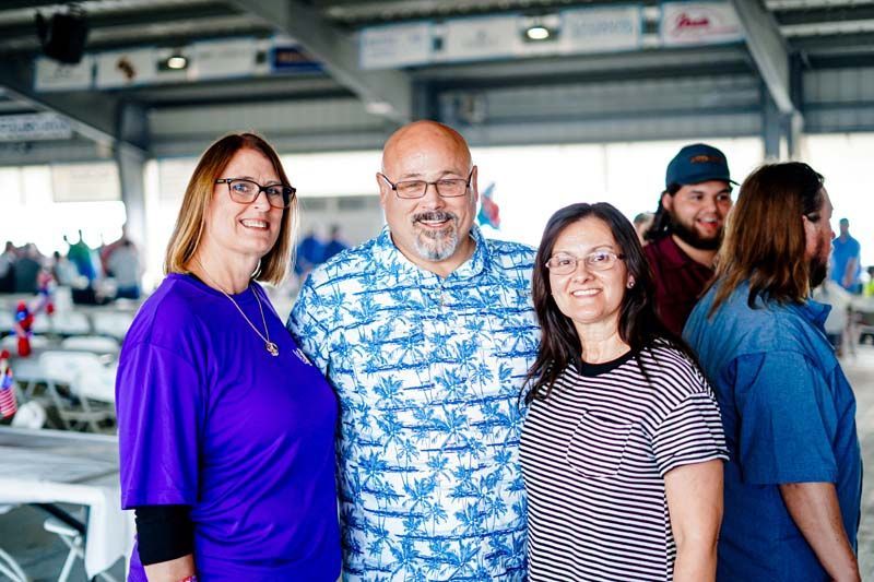 Four people smiling indoors, with a man in a blue patterned shirt centered in front.