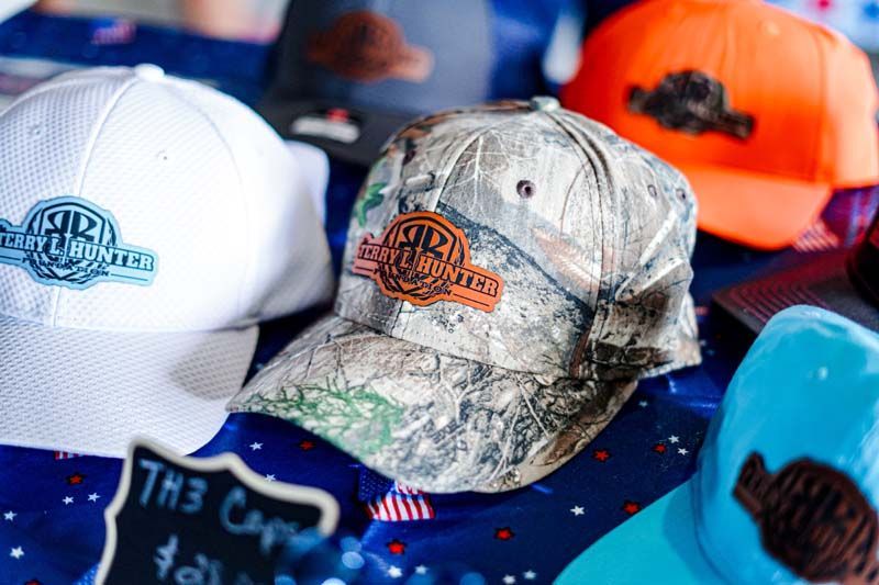 Assorted colorful baseball caps displayed on a table, including white, camo, orange, blue, and teal hats.
