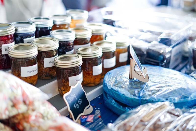 Assorted jars of preserved foods on a table, with blue-wrapped items and small labels in a market setting