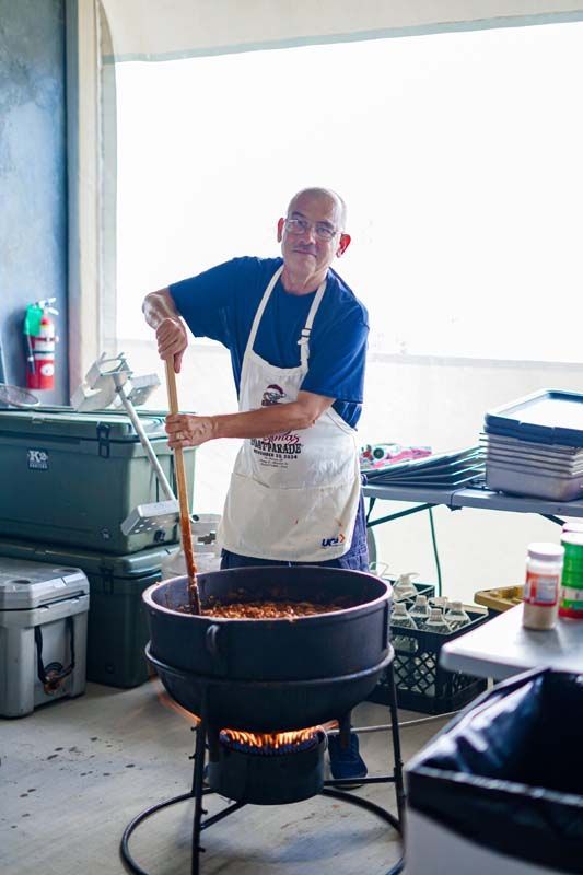 Person stirring a large pot over an open flame in a bright kitchen with stacked trays nearby