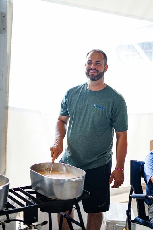 Man stirring a large pot on a stove in a bright kitchen, wearing a green shirt and black shorts.