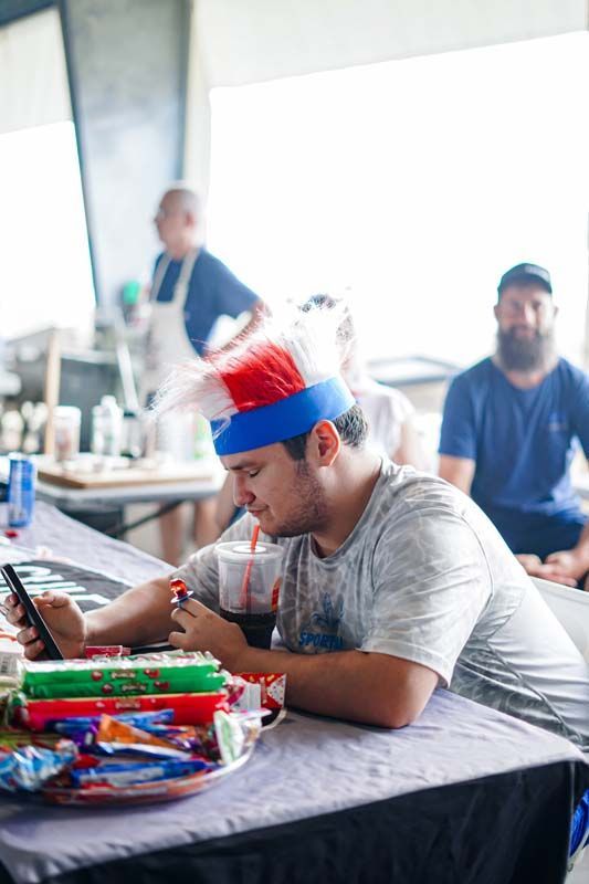 Man in a red-white-blue headband using a phone at a cluttered table indoors, with snacks and drinks nearby