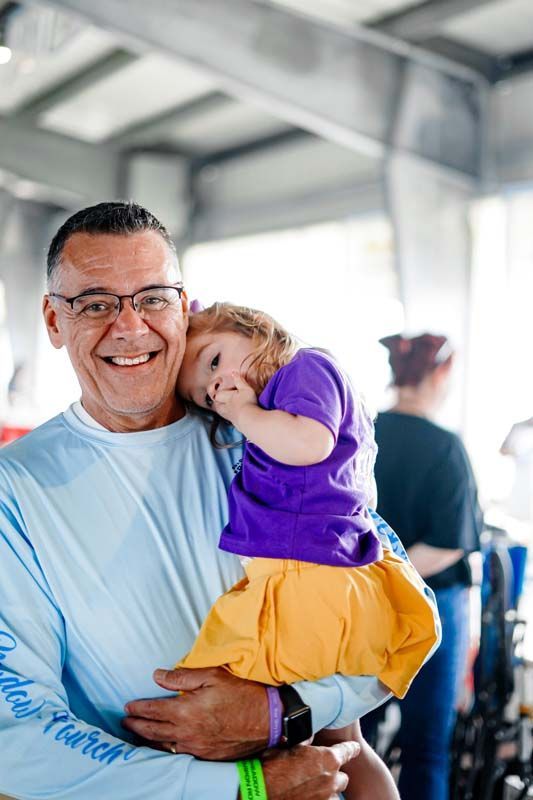 Smiling man holding a child in a purple shirt and yellow skirt indoors under a covered structure
