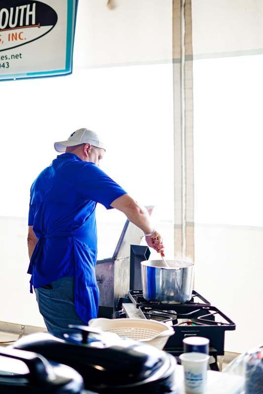 Chef in a blue shirt stirring a large pot on a stove in a bright kitchen