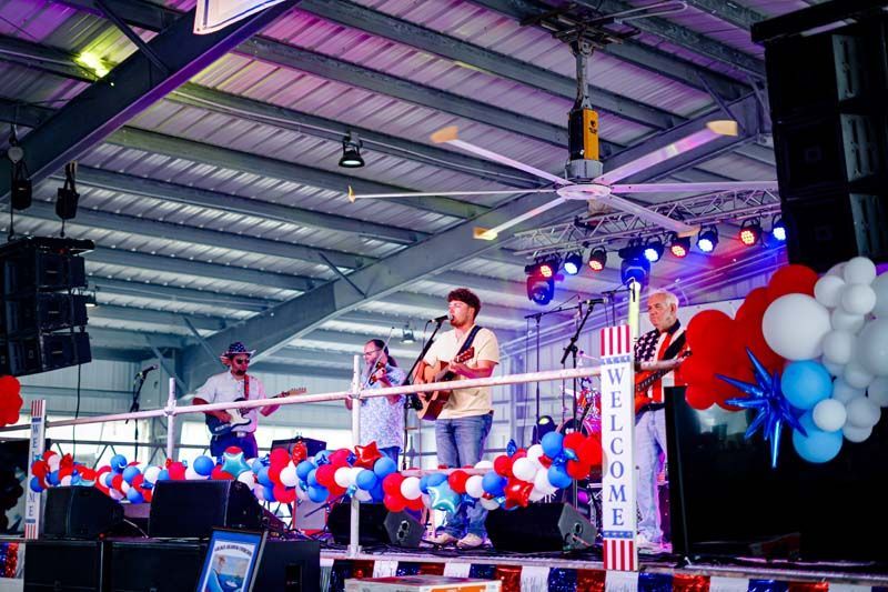 Band performing on a decorated indoor stage with red, white, and blue balloons and lights
