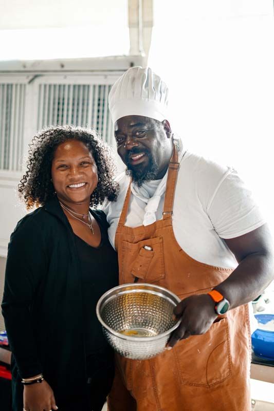 Two smiling people in a kitchen, one holding a metal strainer and wearing a chef hat and apron.