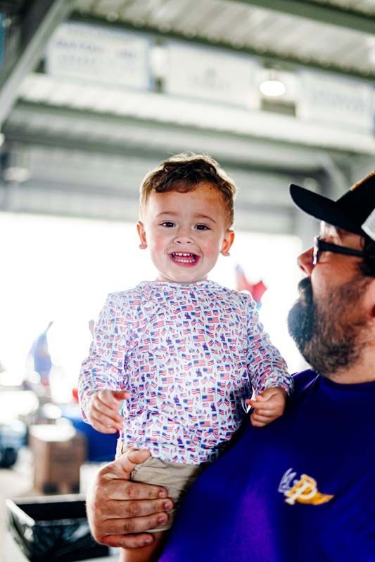 Smiling toddler in patterned shirt being held by a man in a cap indoors