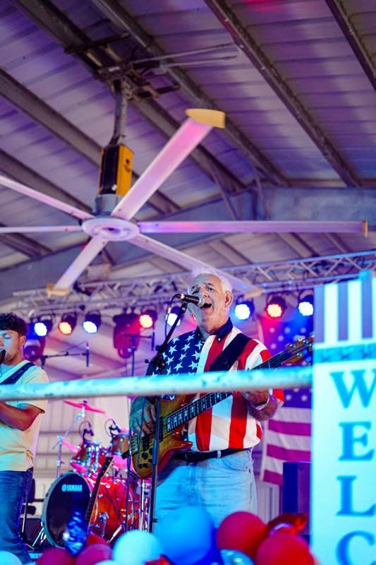 Singer performing on a small stage with colorful lights, red-striped shirt, and American flag background
