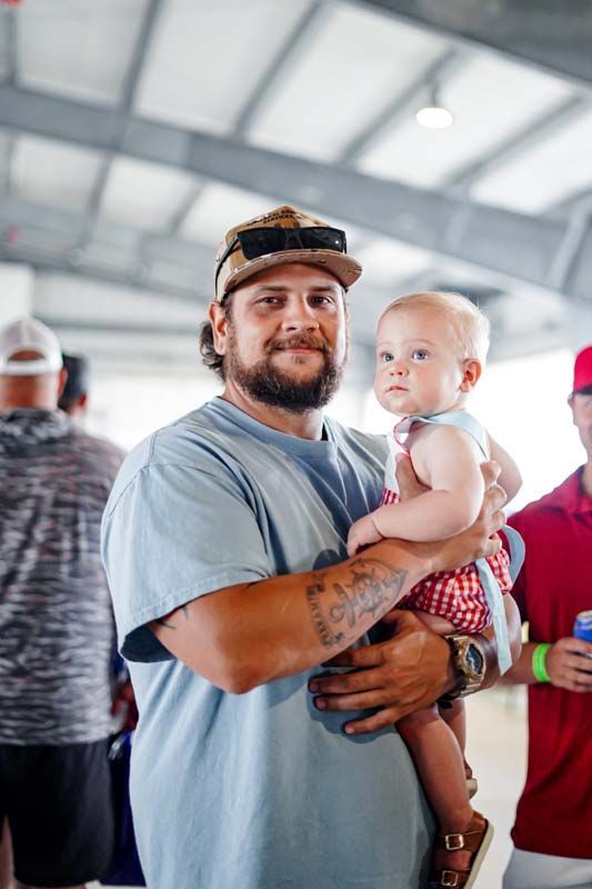 Man holding a baby indoors at a crowded event, both facing the camera.