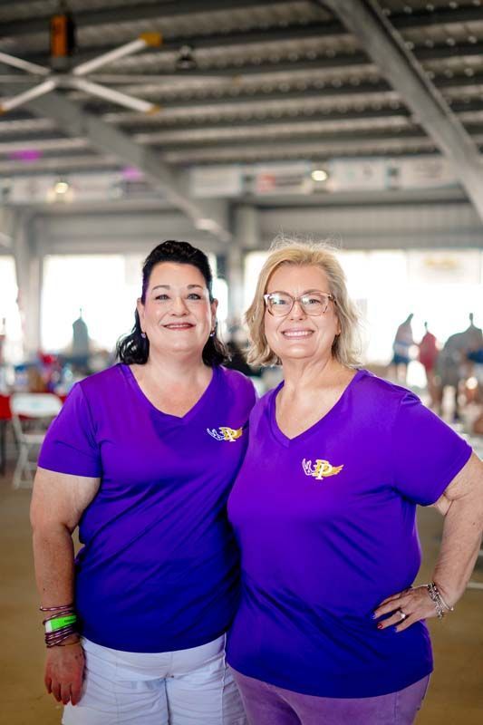 Two women in purple shirts smiling indoors under a metal-roofed pavilion