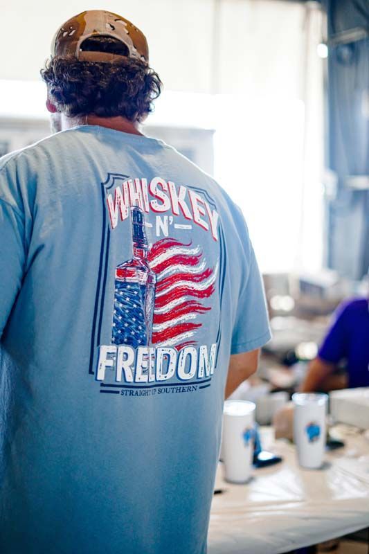 Person wearing a blue “Whiskey and Freedom” shirt stands in a bright room near a table with cups.