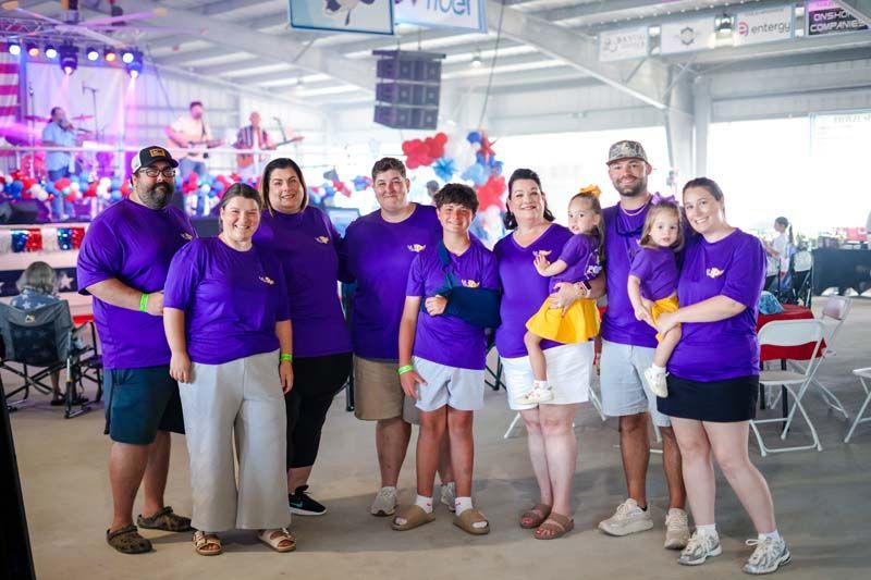 Group of people in purple shirts posing indoors at a community event, some holding purple and gold balloons