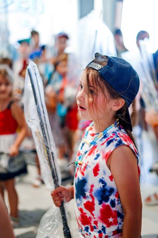 A little girl in a tie dye dress and a baseball cap is holding a flag.