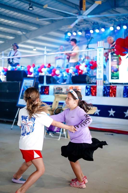 Two girls dancing at a patriotic indoor event with a stage and flags in the background