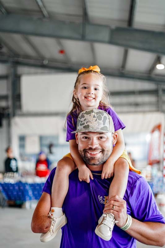 Smiling child on a man's shoulders in a gym, both wearing purple shirts