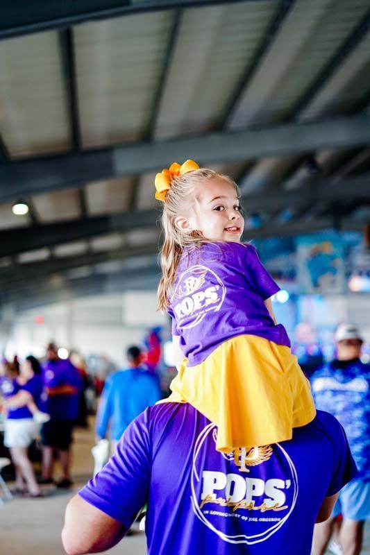 Child in purple and yellow outfit riding on an adult’s shoulders at an indoor sports venue