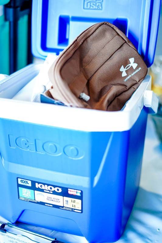 Blue Igloo cooler with a brown Under Armour bag inside, sitting open on a white surface.